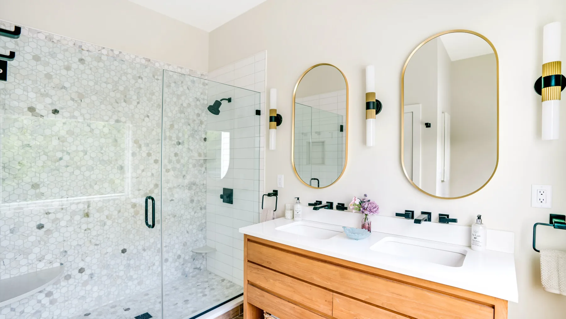 A bright bathroom with a glass shower, double sink, and oval mirrors.