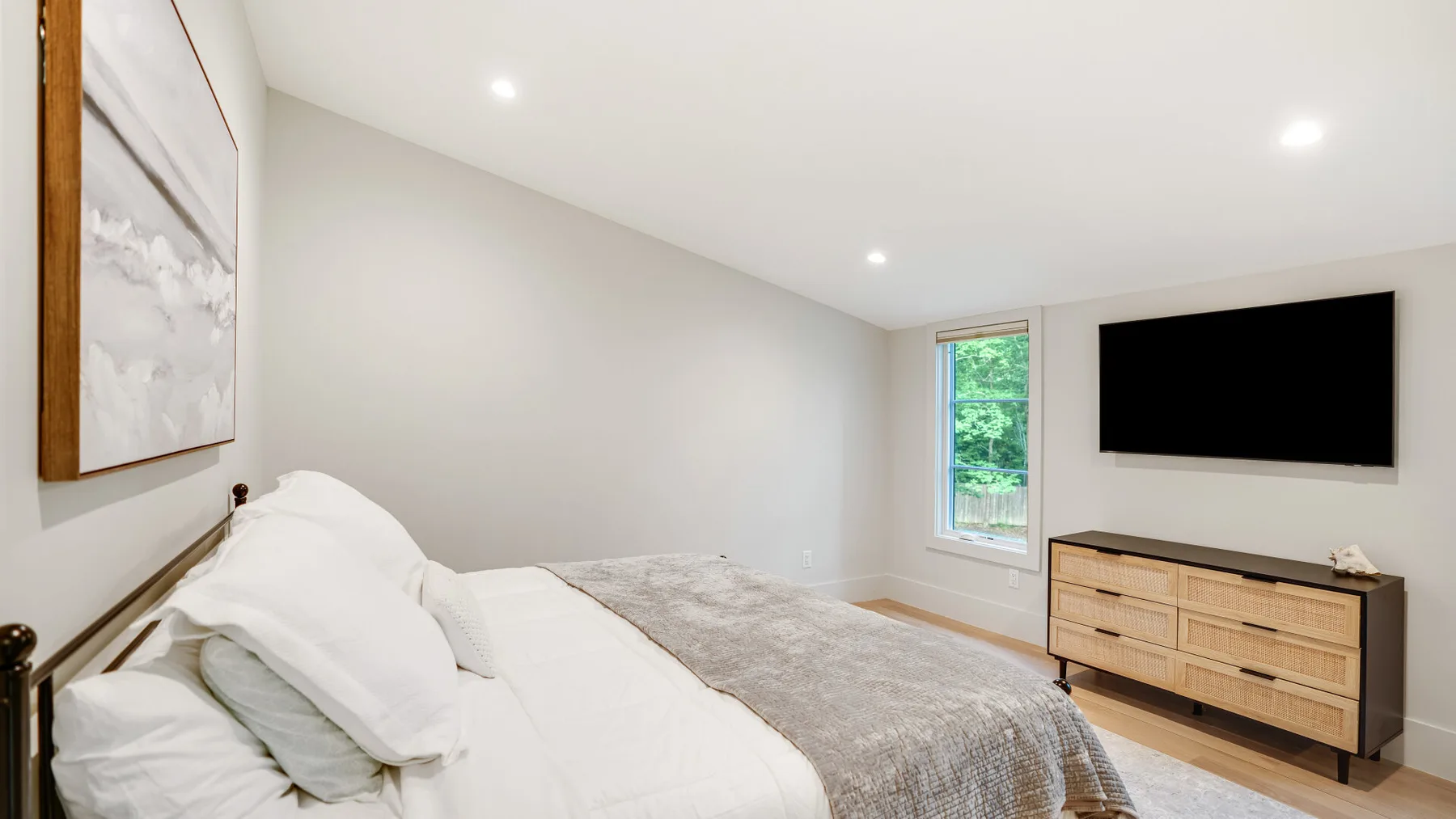 A bright bedroom with a bed, a wall-mounted television, and a window.