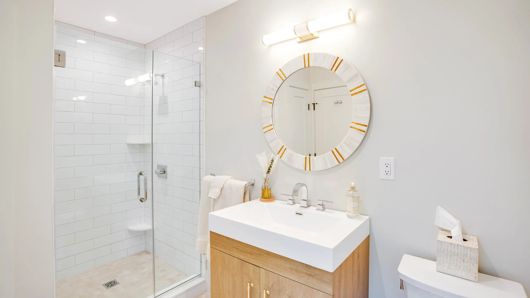 A bright bathroom with a glass shower and a sink vanity beneath a round mirror.