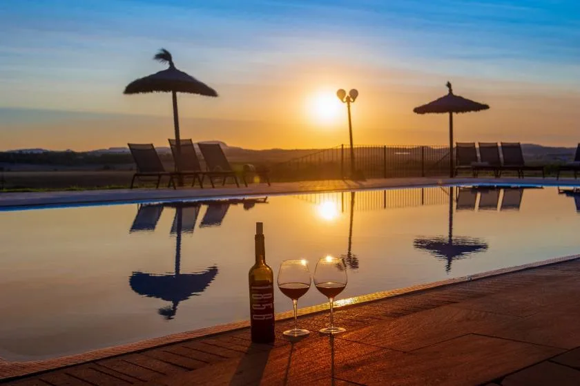 An outdoor pool reflects the sunset, with lounge seating and a pair of glasses nearby.