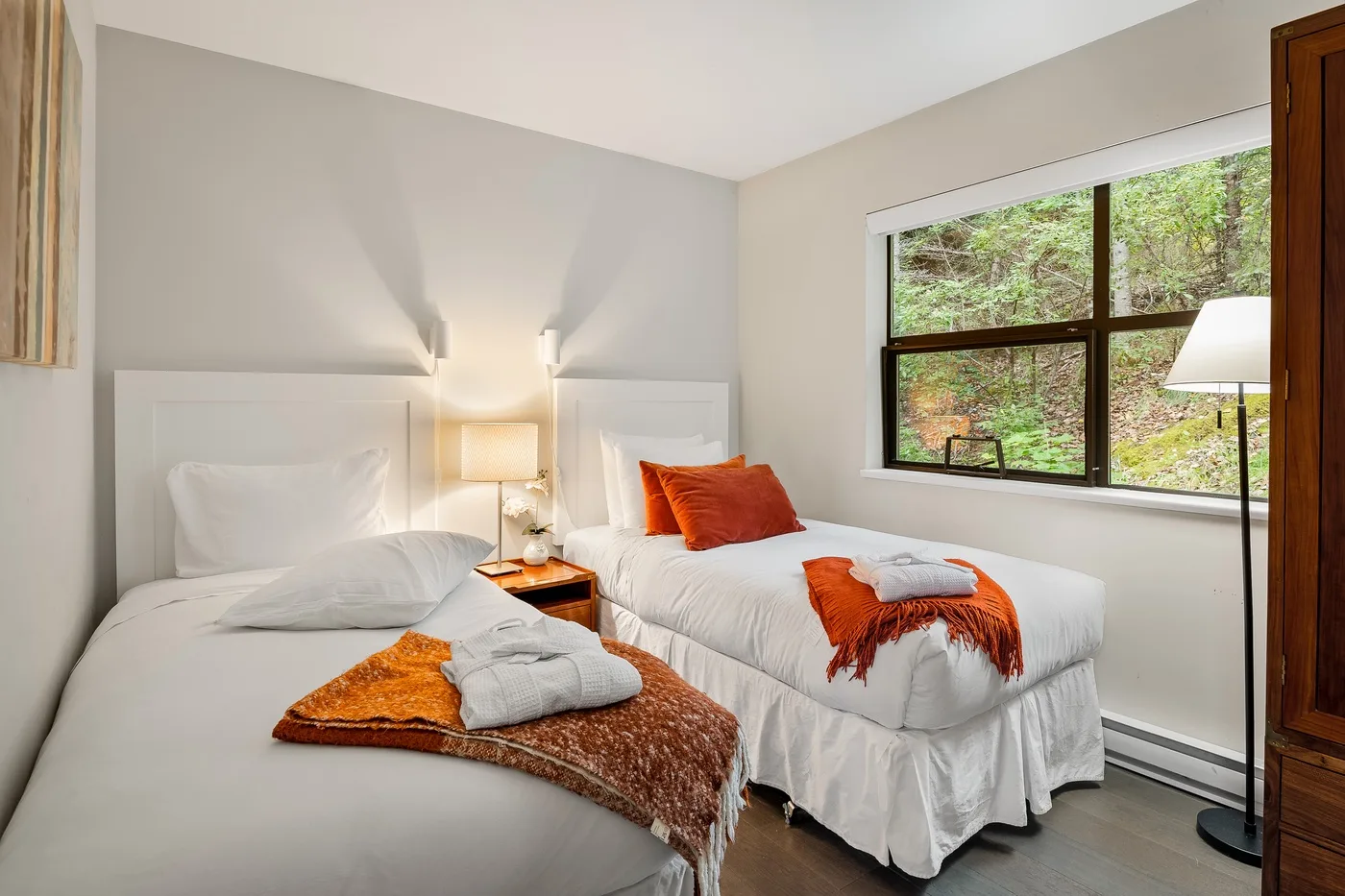 Un dormitorio con dos camas, detalles naranjas y una ventana amplia con vista a la vegetación.