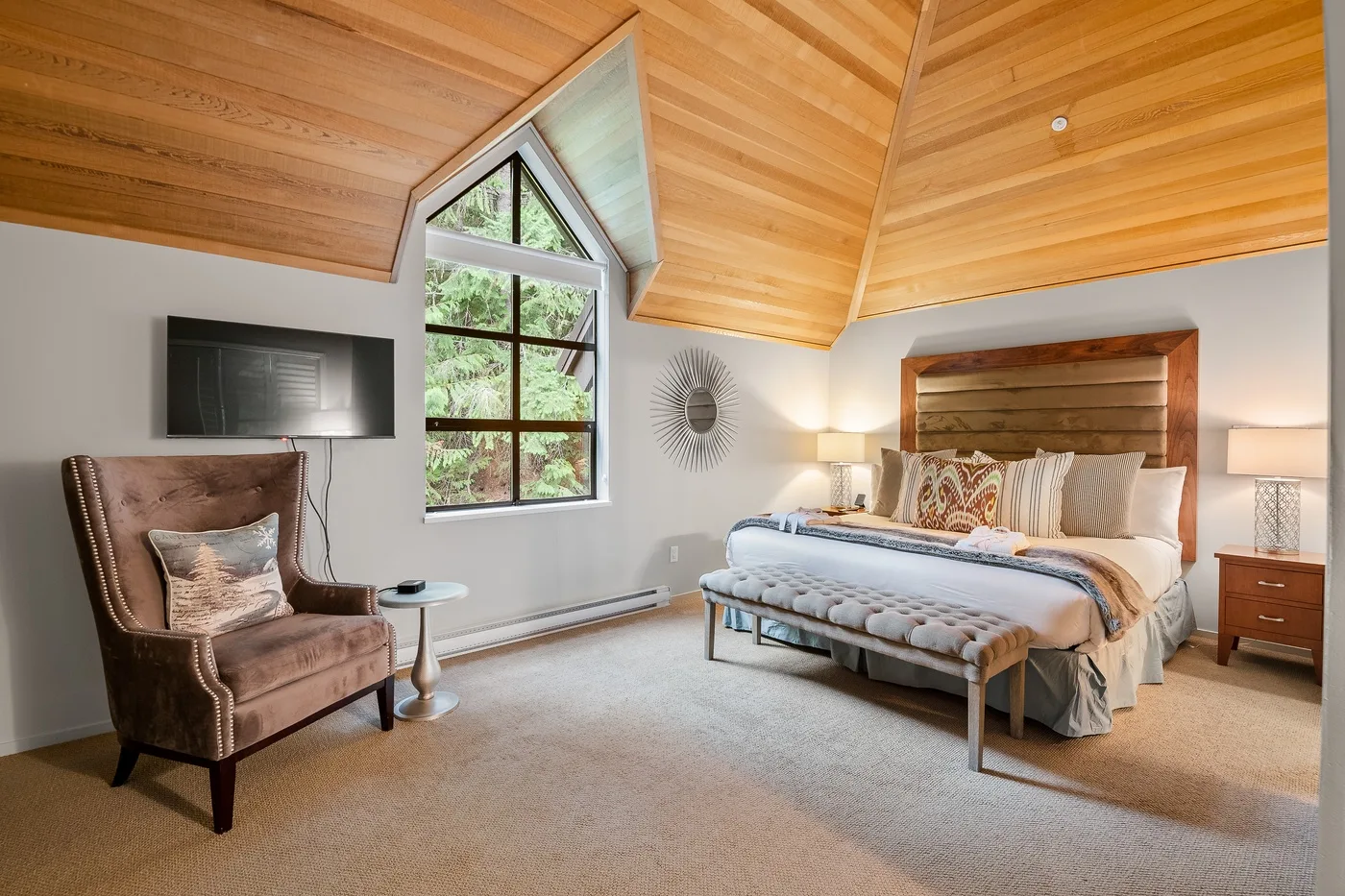 Un dormitorio con una cama, un sillón y una ventana grande bajo un techo inclinado de madera.