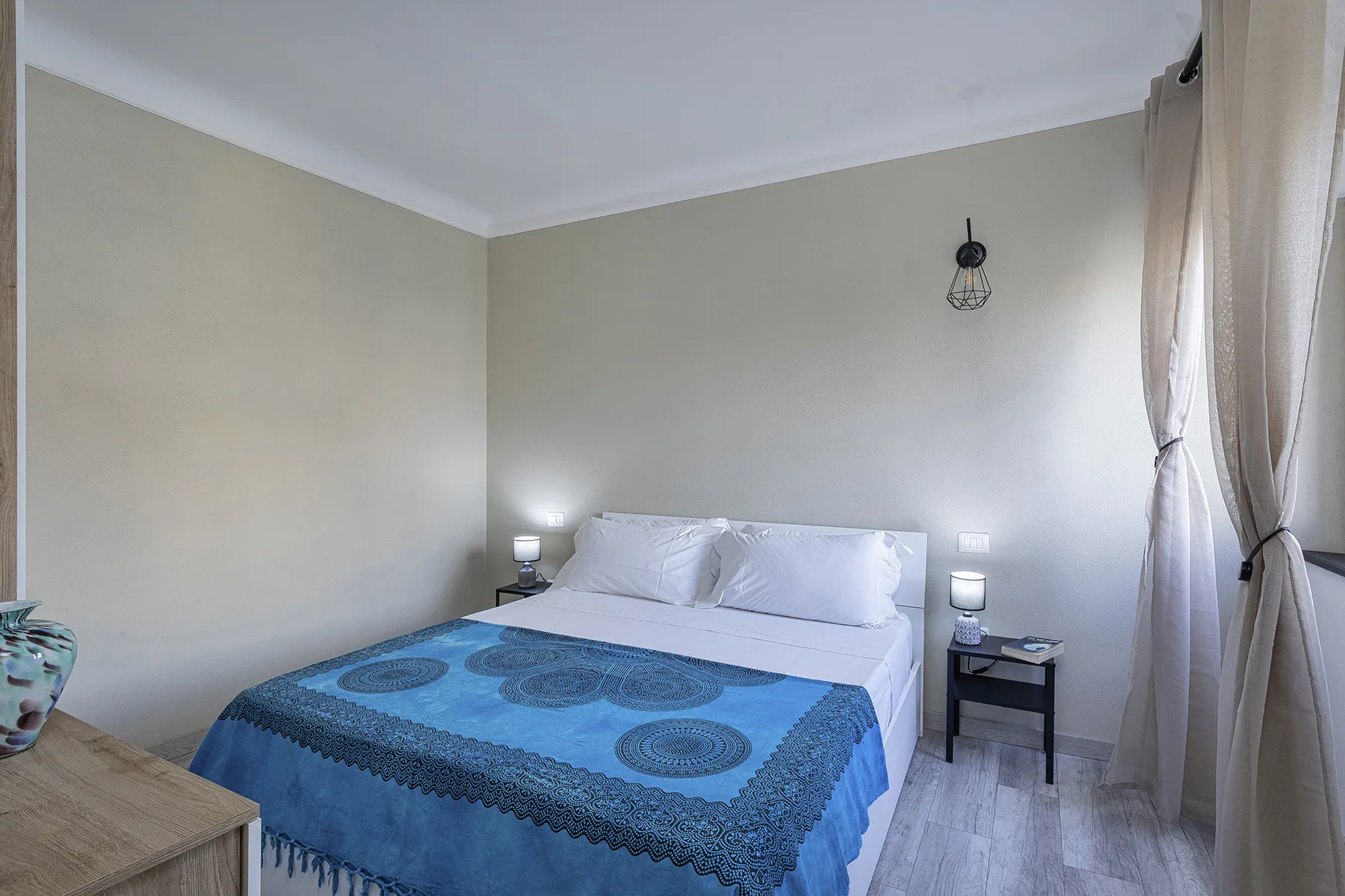 A bedroom with a bed covered by a blue patterned bedspread and curtains by the window.
