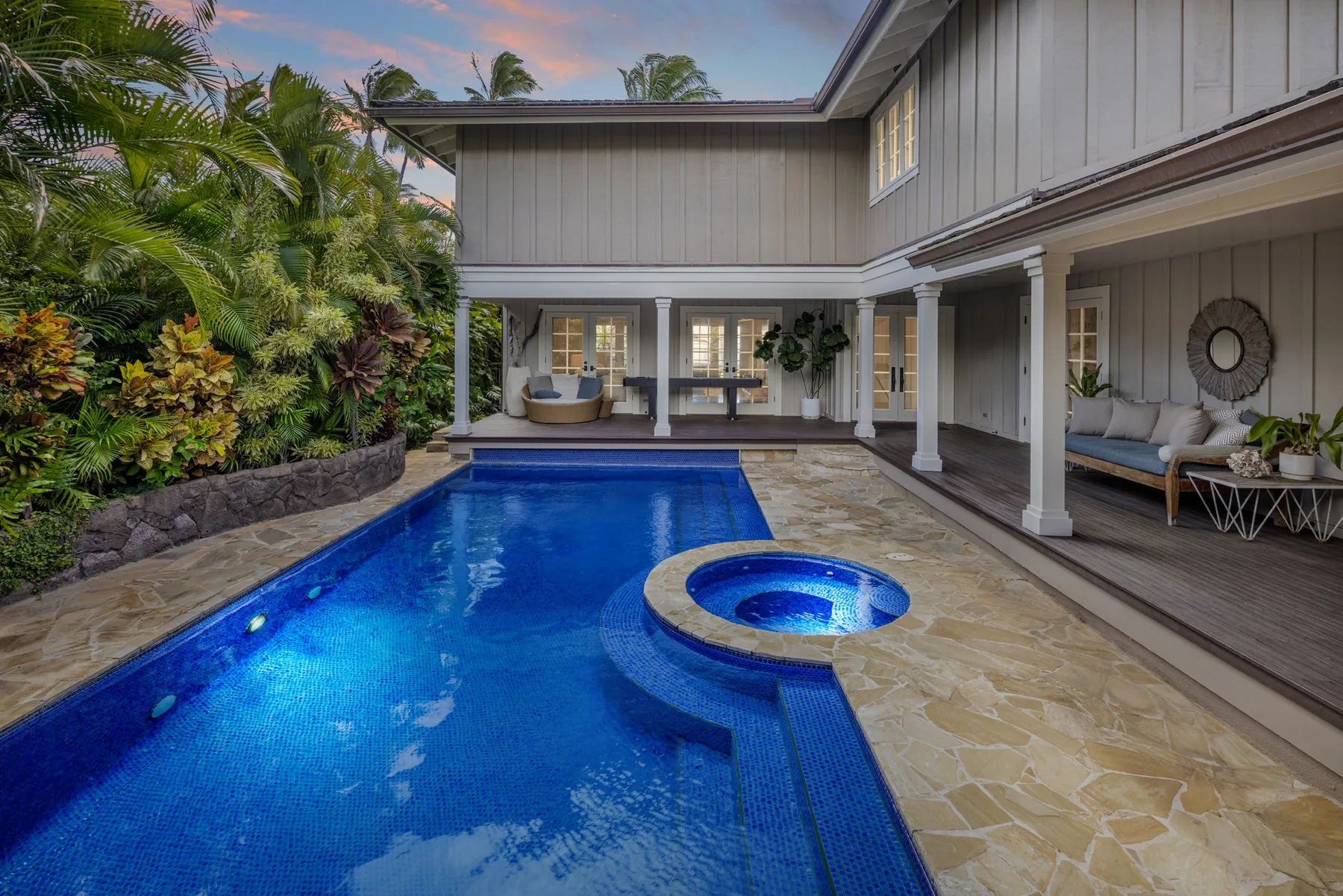 A backyard pool beside a covered patio with seating and tropical plants.