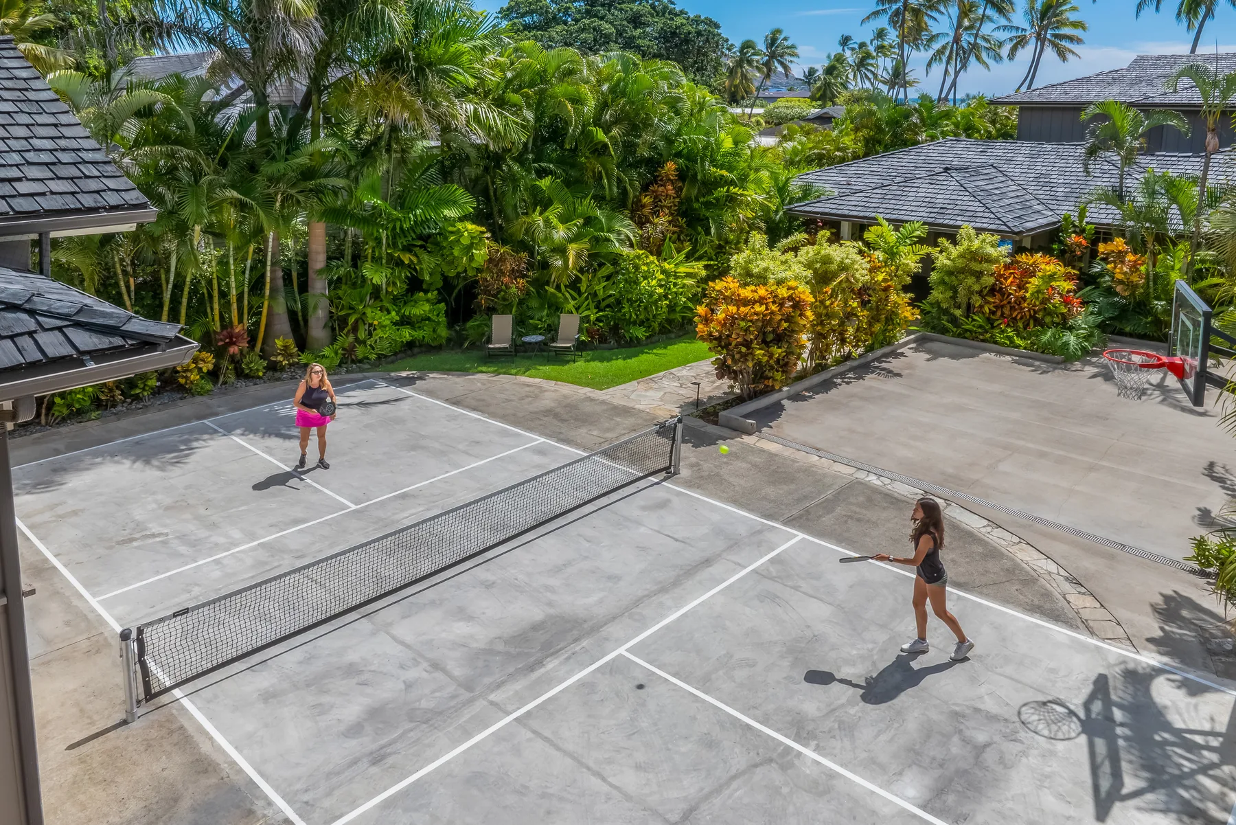 Outdoor tennis court with two players and lush tropical landscaping.