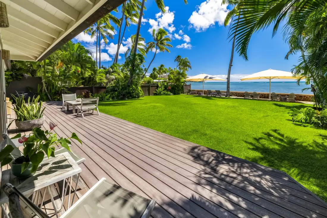 A sunny beachfront garden with a wooden deck, outdoor seating, and ocean views.