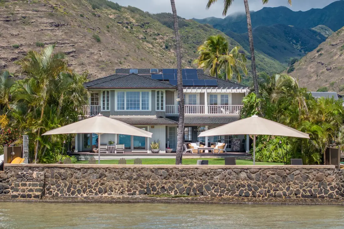 A two-story house with shaded patio seating stands among palm trees against a mountain backdrop.