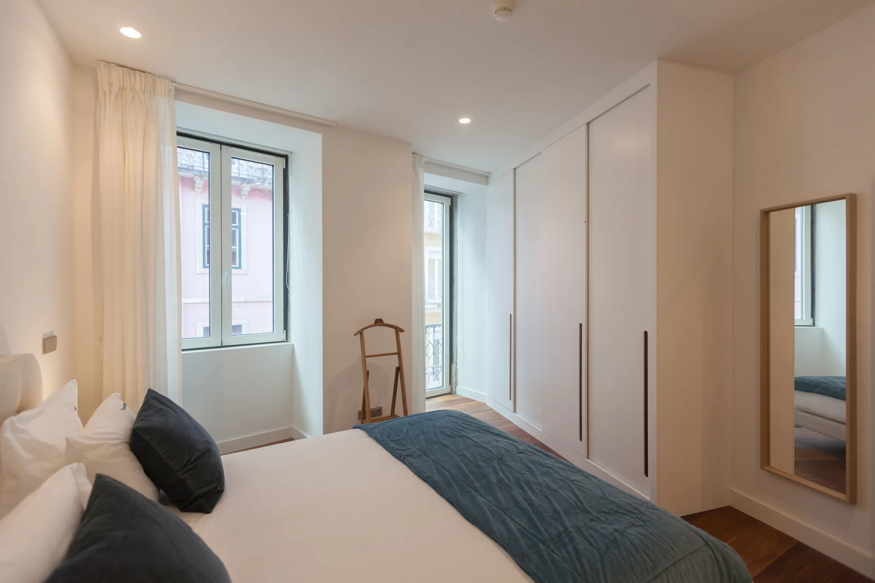 Un dormitorio con una cama, ropa de cama oscura y ventanas altas.
