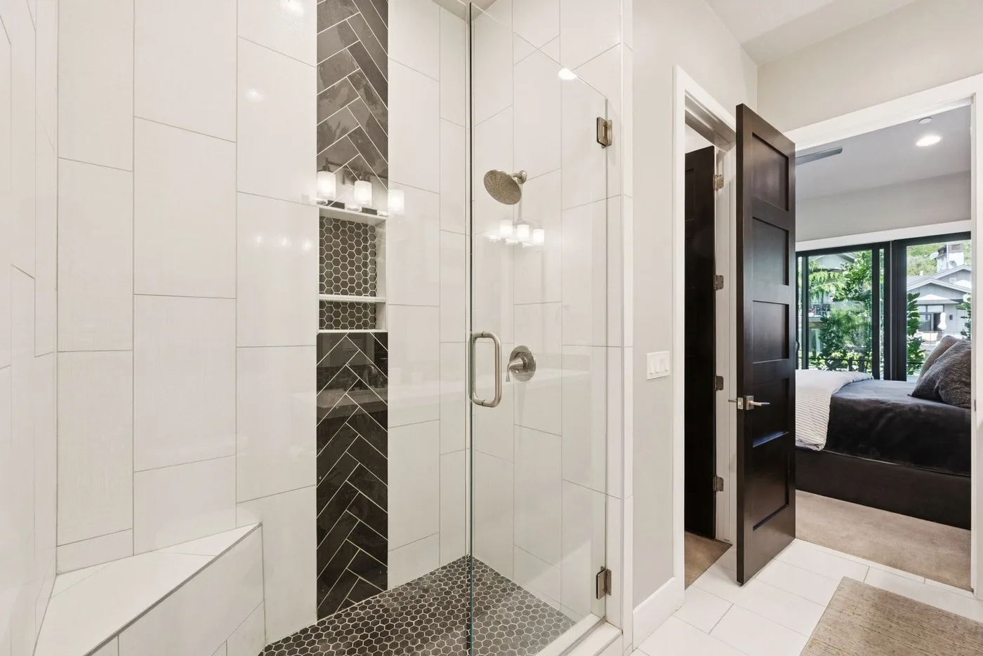 A tiled bathroom with a glass shower and an open doorway to a bedroom.