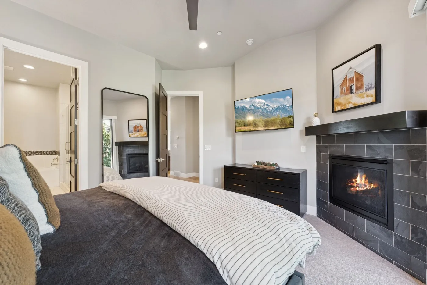 A bedroom with a bed, a dark fireplace, and framed landscape artwork.