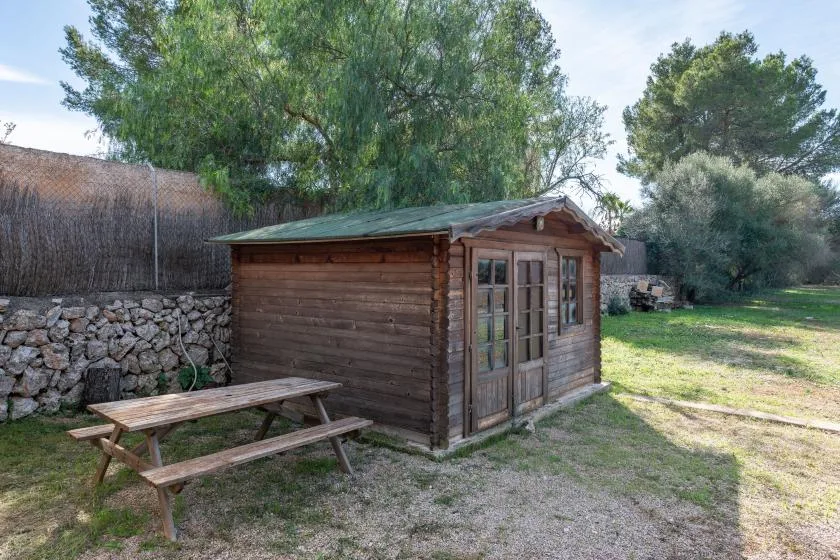 Un pequeño cobertizo de madera está junto a una mesa de picnic bajo árboles frondosos.