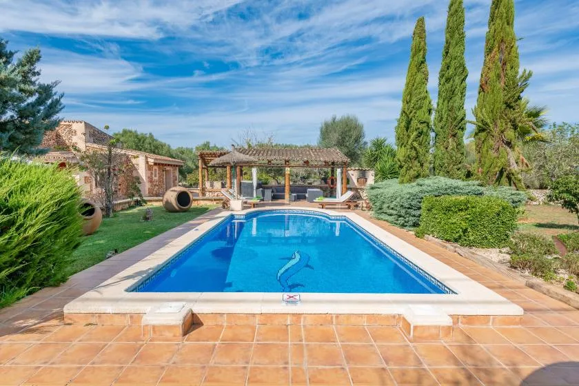 Una piscina al aire libre está junto a una pérgola sombreada y un jardín ajardinado.