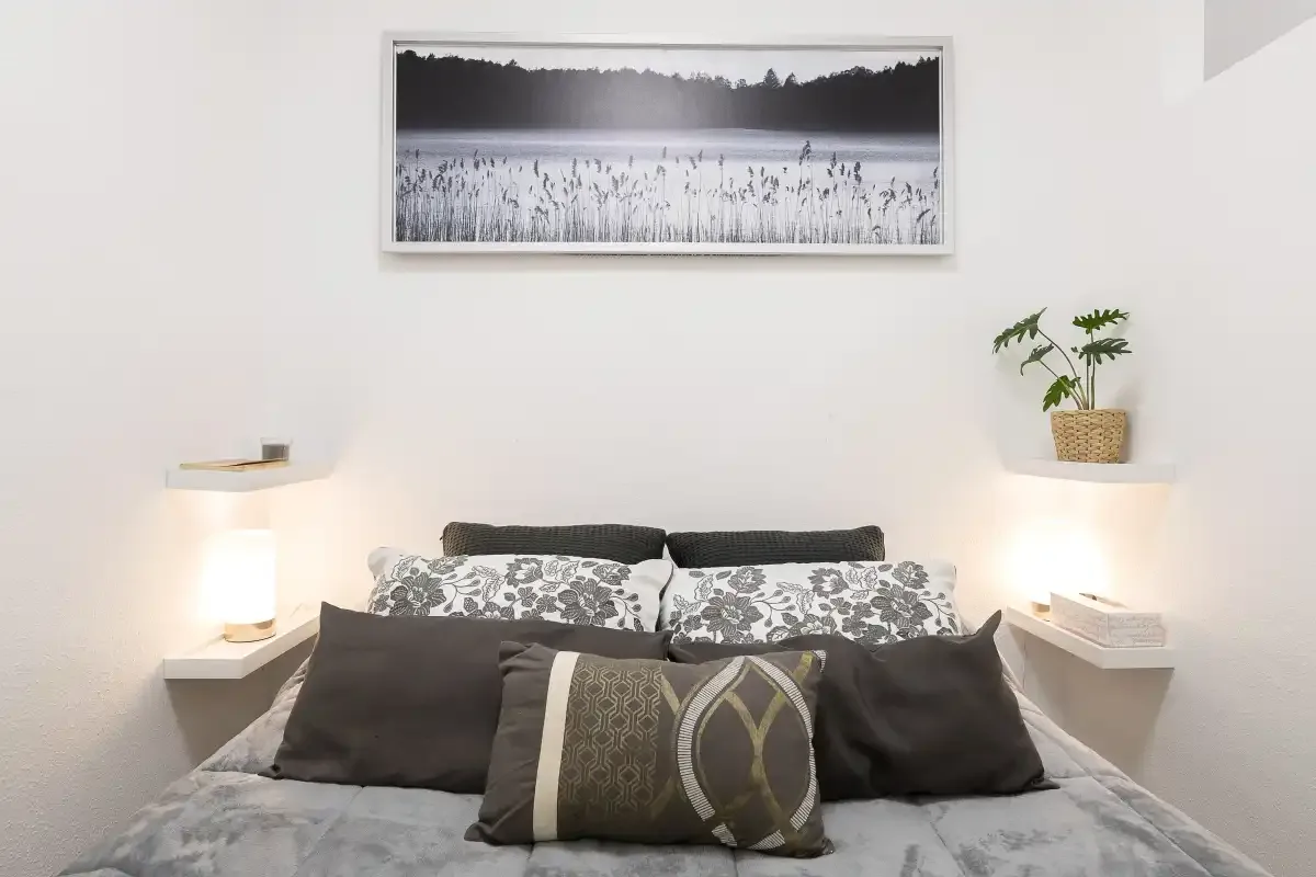 A bedroom with a neatly made bed, patterned pillows, and a framed landscape above it.