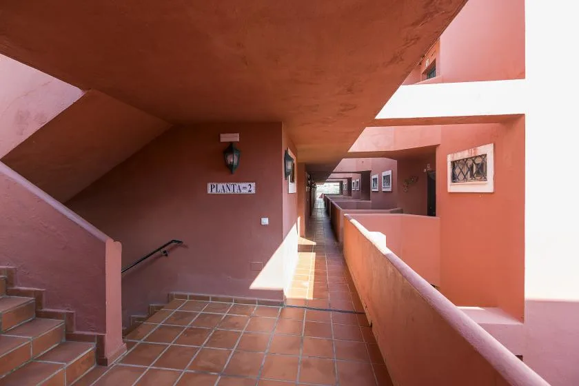 A tiled outdoor corridor runs past terracotta walls and a stairway.