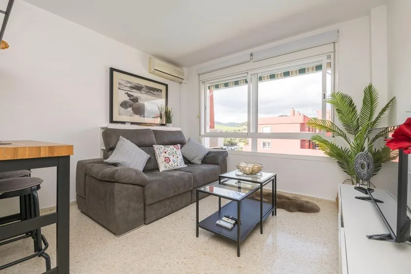 Una sala luminosa con asientos grises, una mesa de centro de vidrio y ventanas grandes.