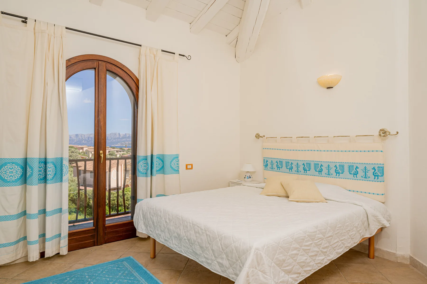 Un dormitorio luminoso con una cama doble y una puerta acristalada arqueada que da a un balcón.