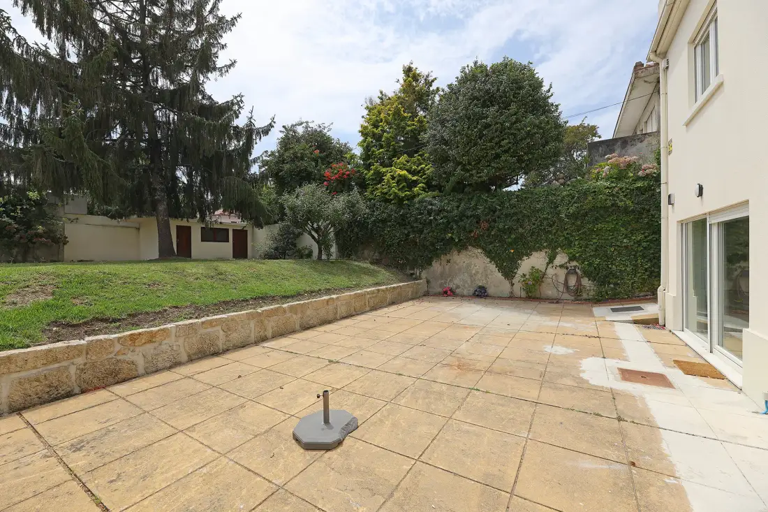 A paved outdoor patio borders a grassy garden with mature trees and greenery.