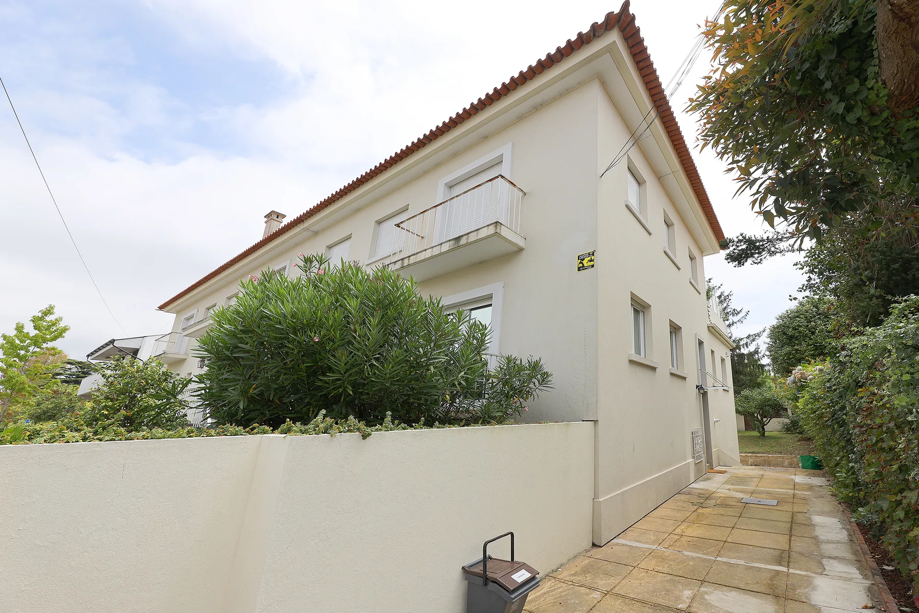 A pale two-story house with a paved path beside leafy shrubs.