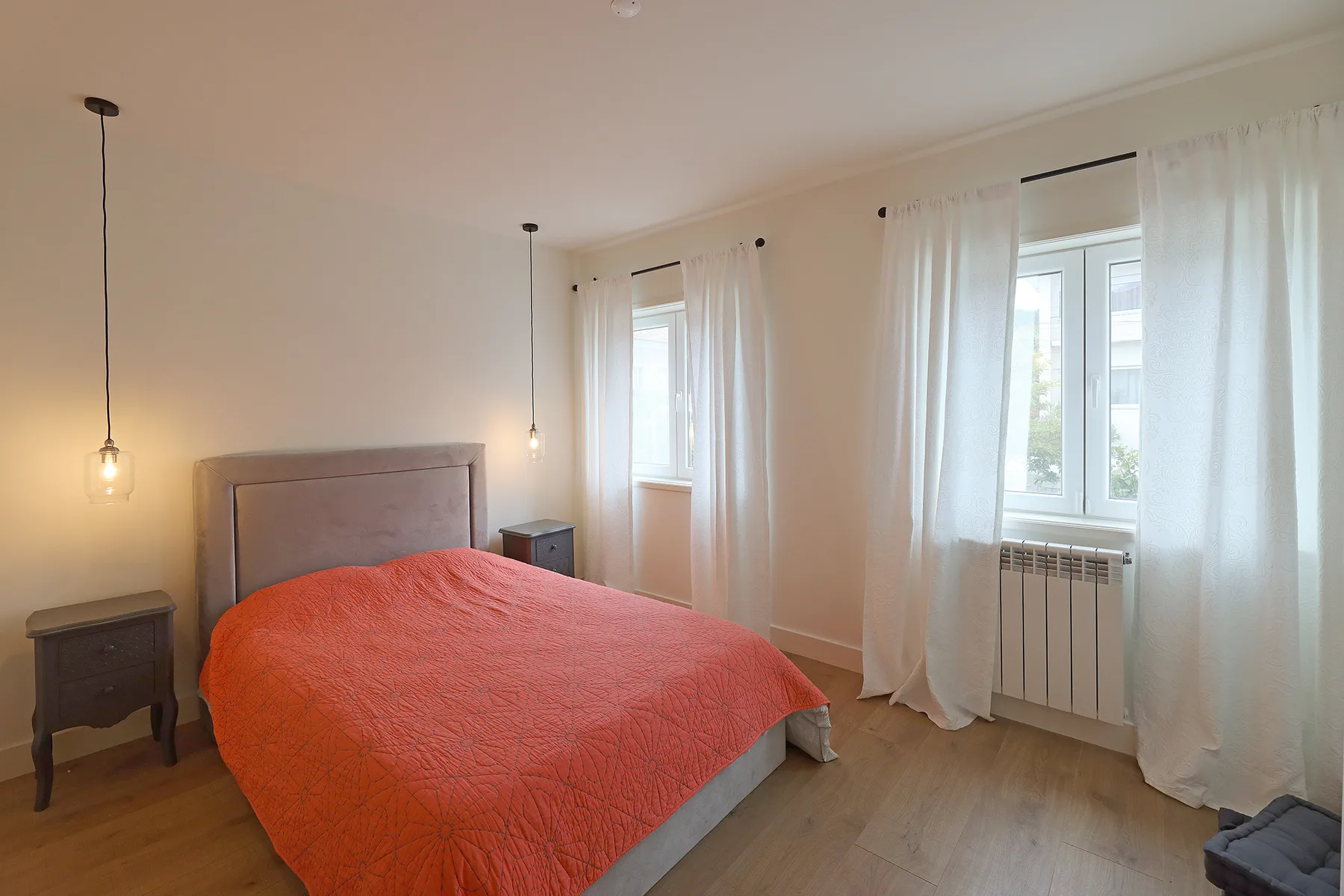A bedroom with a bed covered in an orange quilt and windows with white curtains.