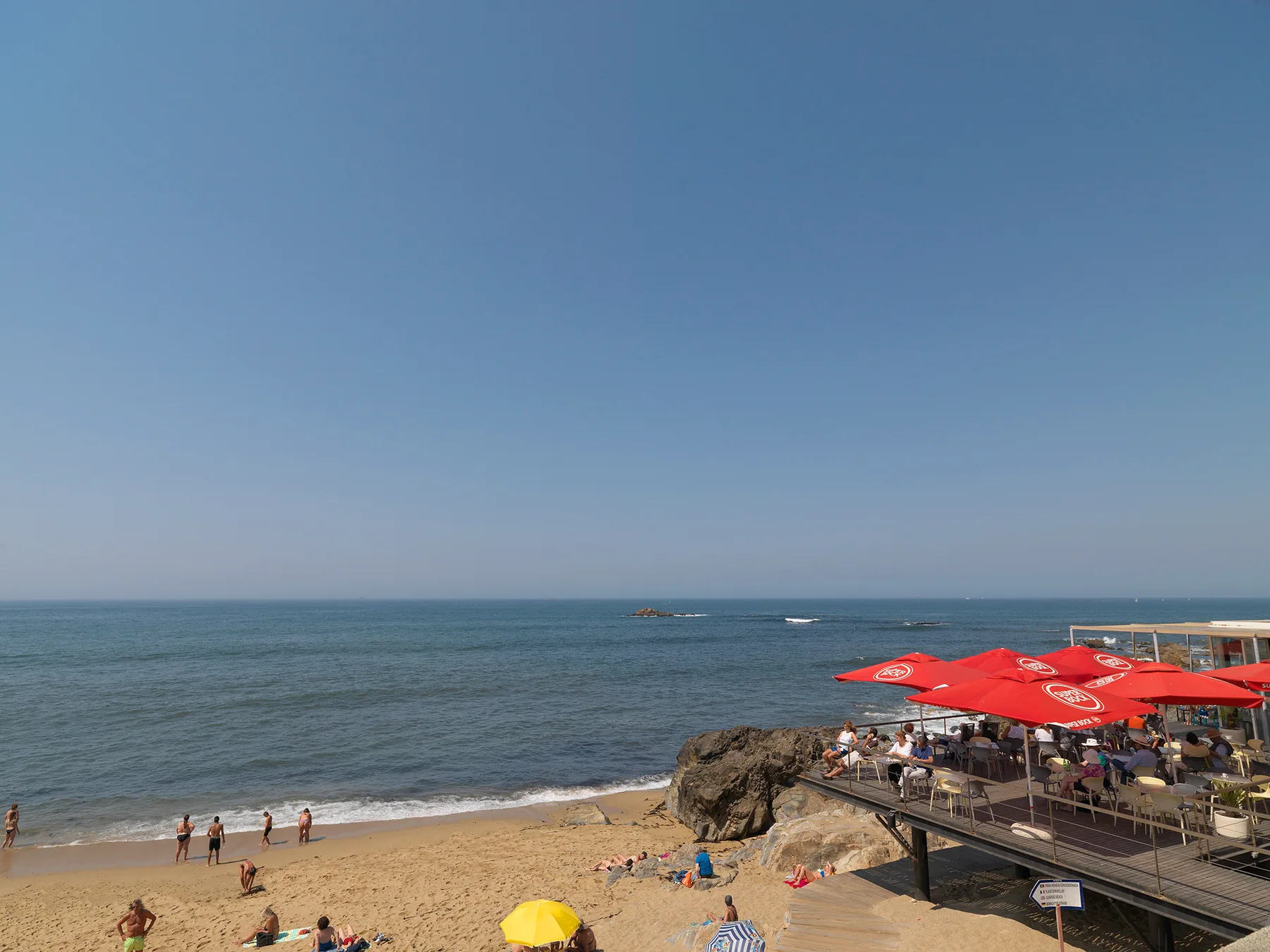 A sandy beach with swimmers and a seaside café beneath a clear sky.