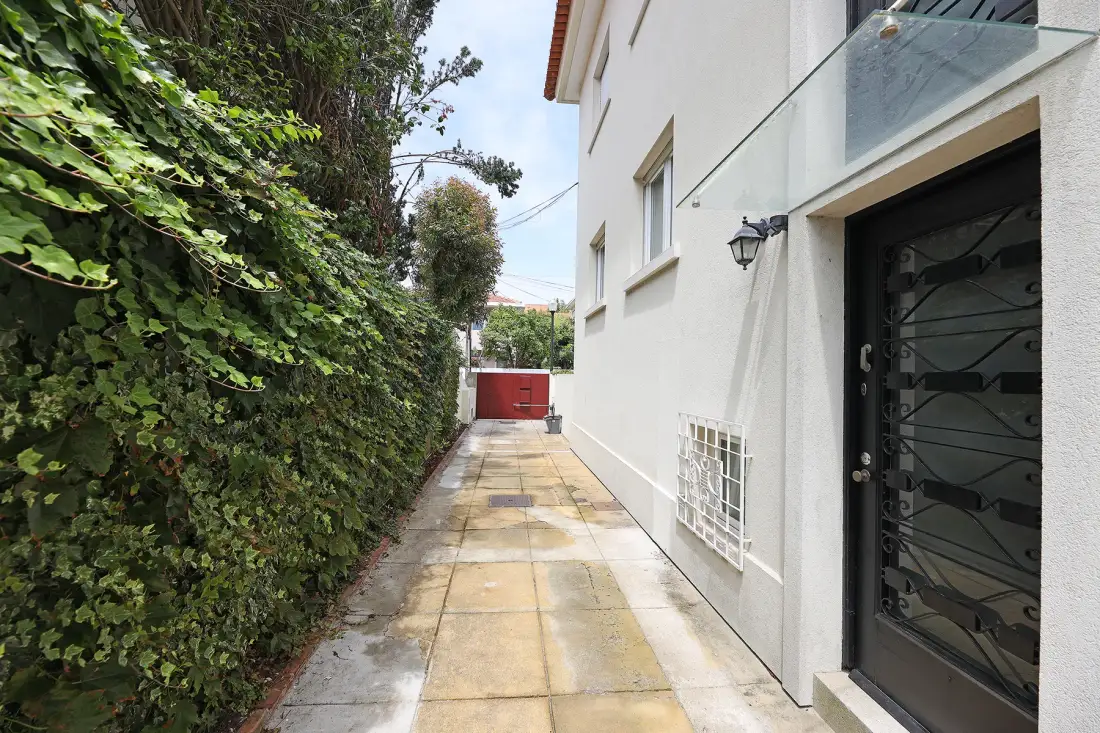 A paved walkway runs alongside a leafy hedge and a white building.