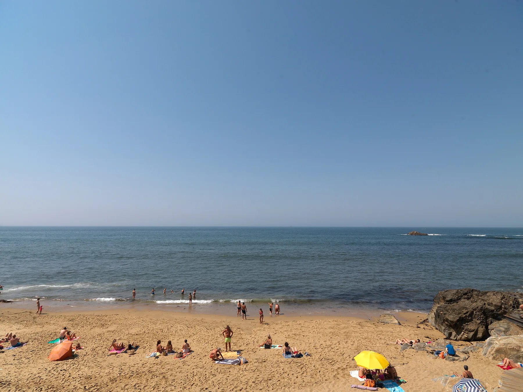 A sandy beach with people relaxing by the water and a rocky outcrop.