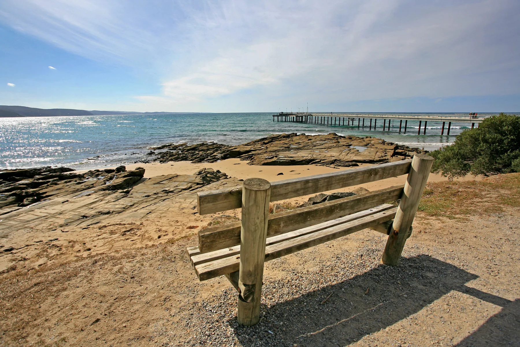 A coastal shoreline with rocky outcrops, calm water, and a wooden bench overlooking the sea.