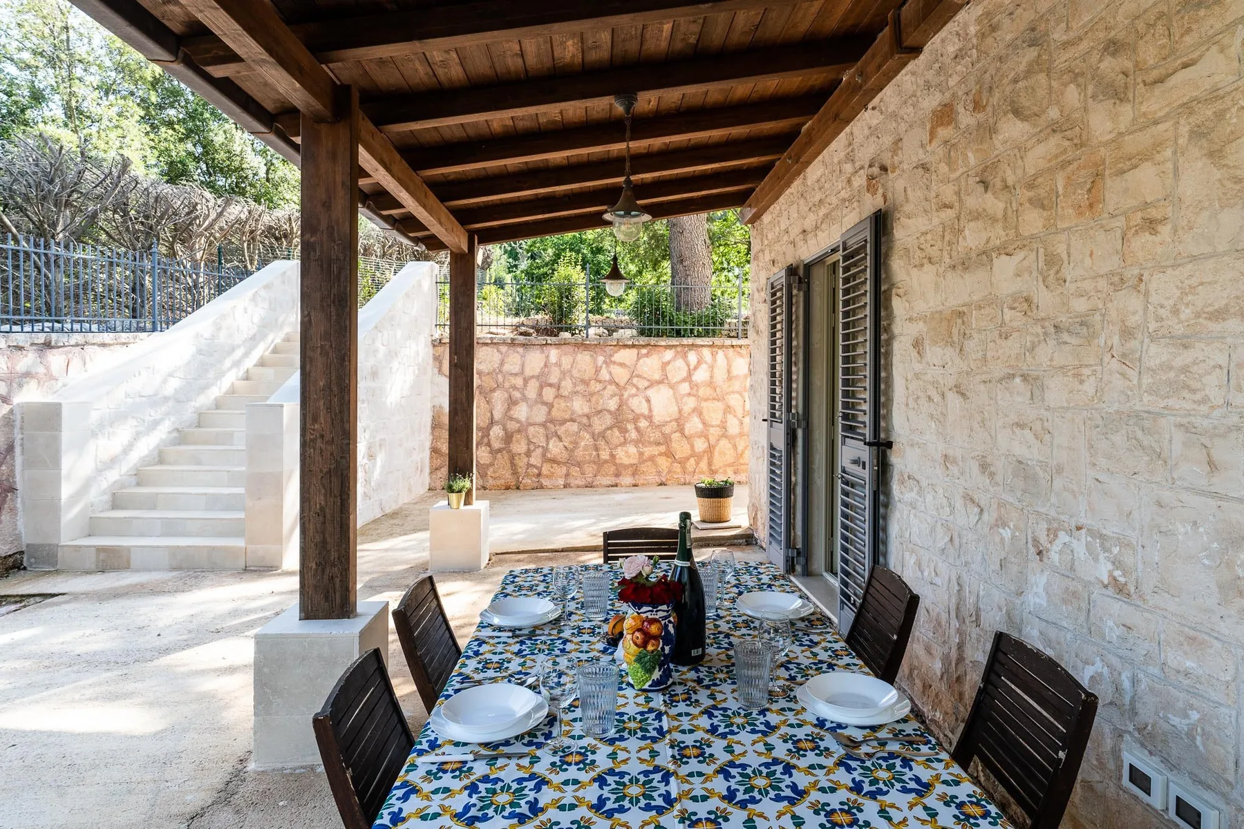 A covered outdoor dining area with a long table beside a stone wall.