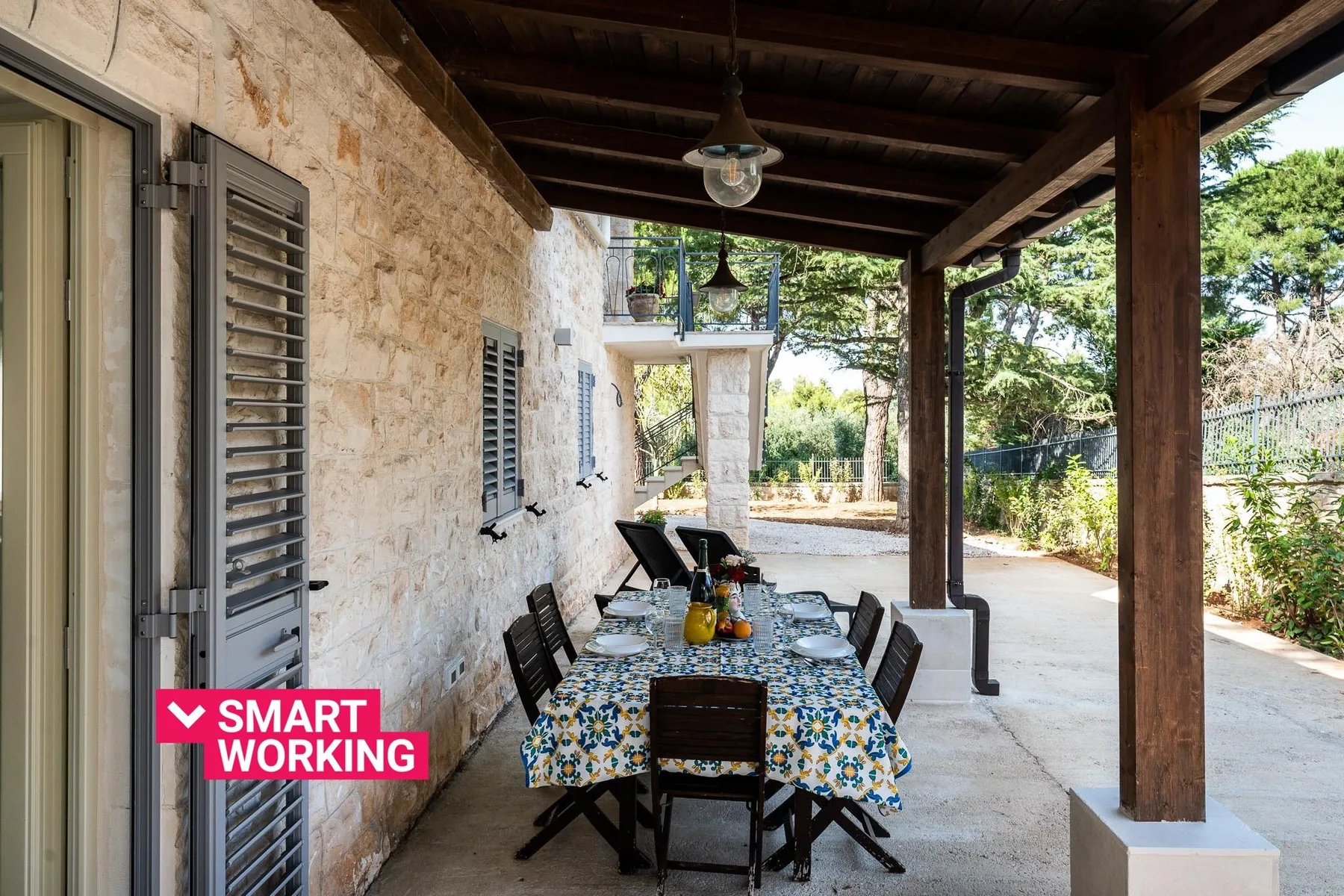 A covered outdoor dining area has a long table set beside a stone wall.