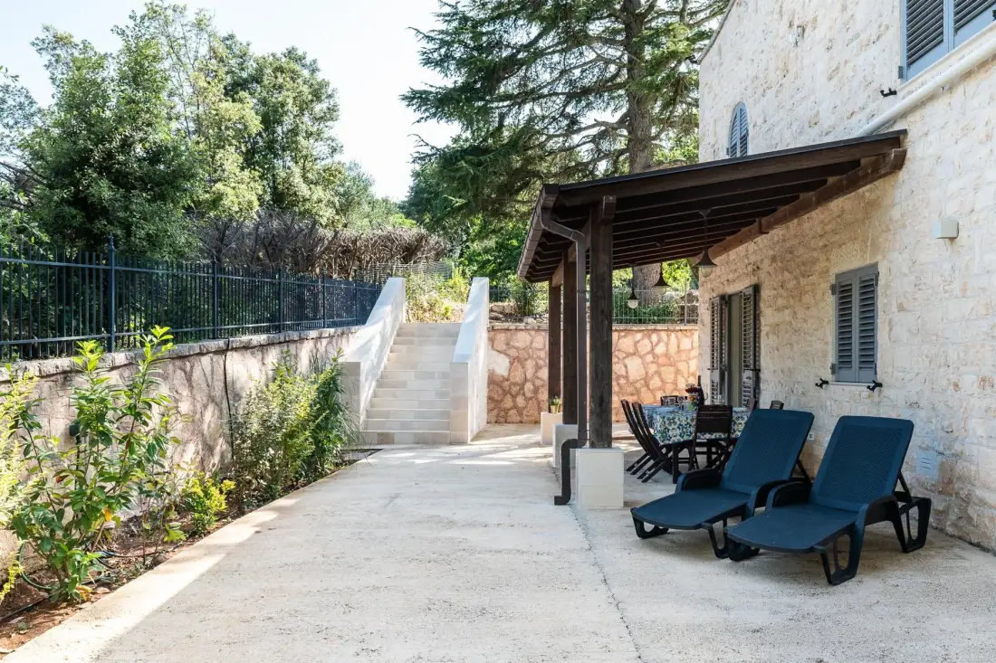 A paved outdoor patio beside a stone house, with seating under a wooden pergola.