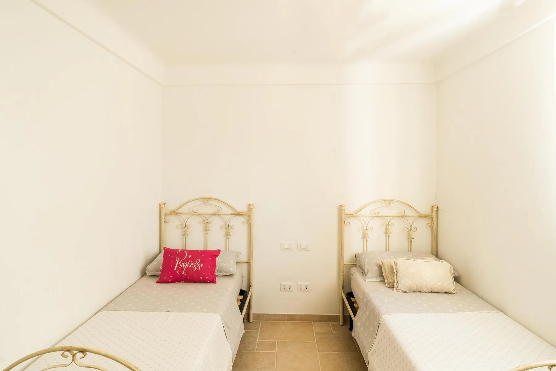 A bright bedroom has two single beds with pale bedding and decorative metal headboards.