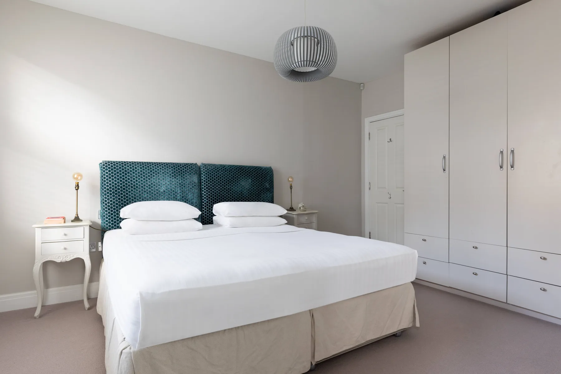 A bedroom with a neatly made bed, teal upholstered headboard, and white wardrobes.