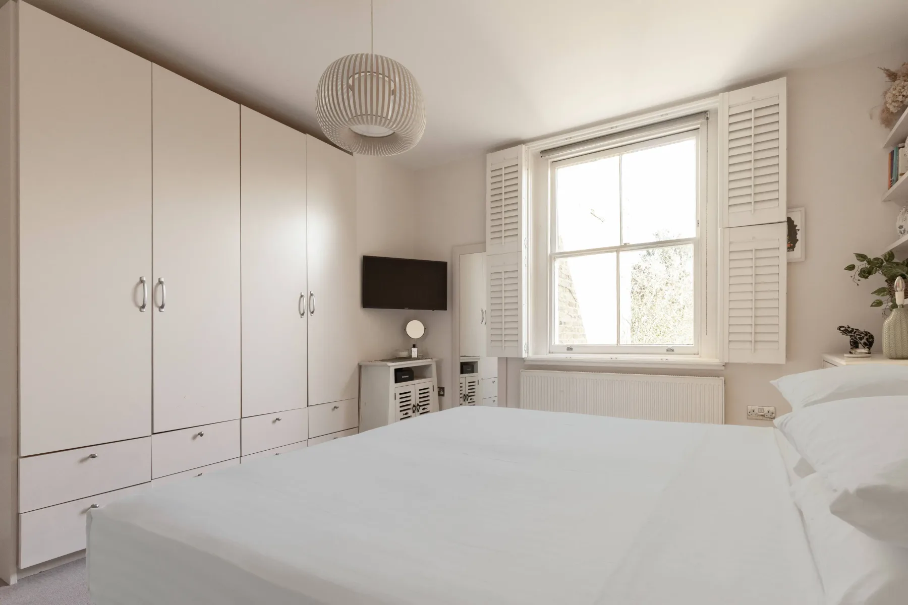 A bright bedroom with a bed, built-in cupboards, and a large window.