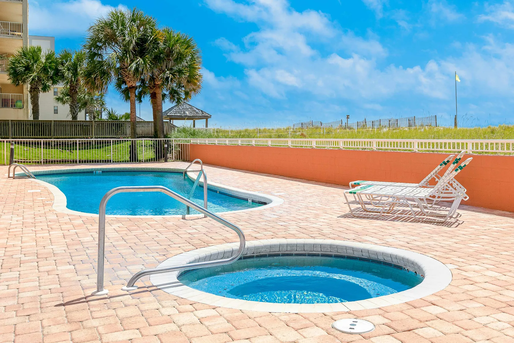 An outdoor pool and circular spa sit on a paved deck beneath a bright sky.