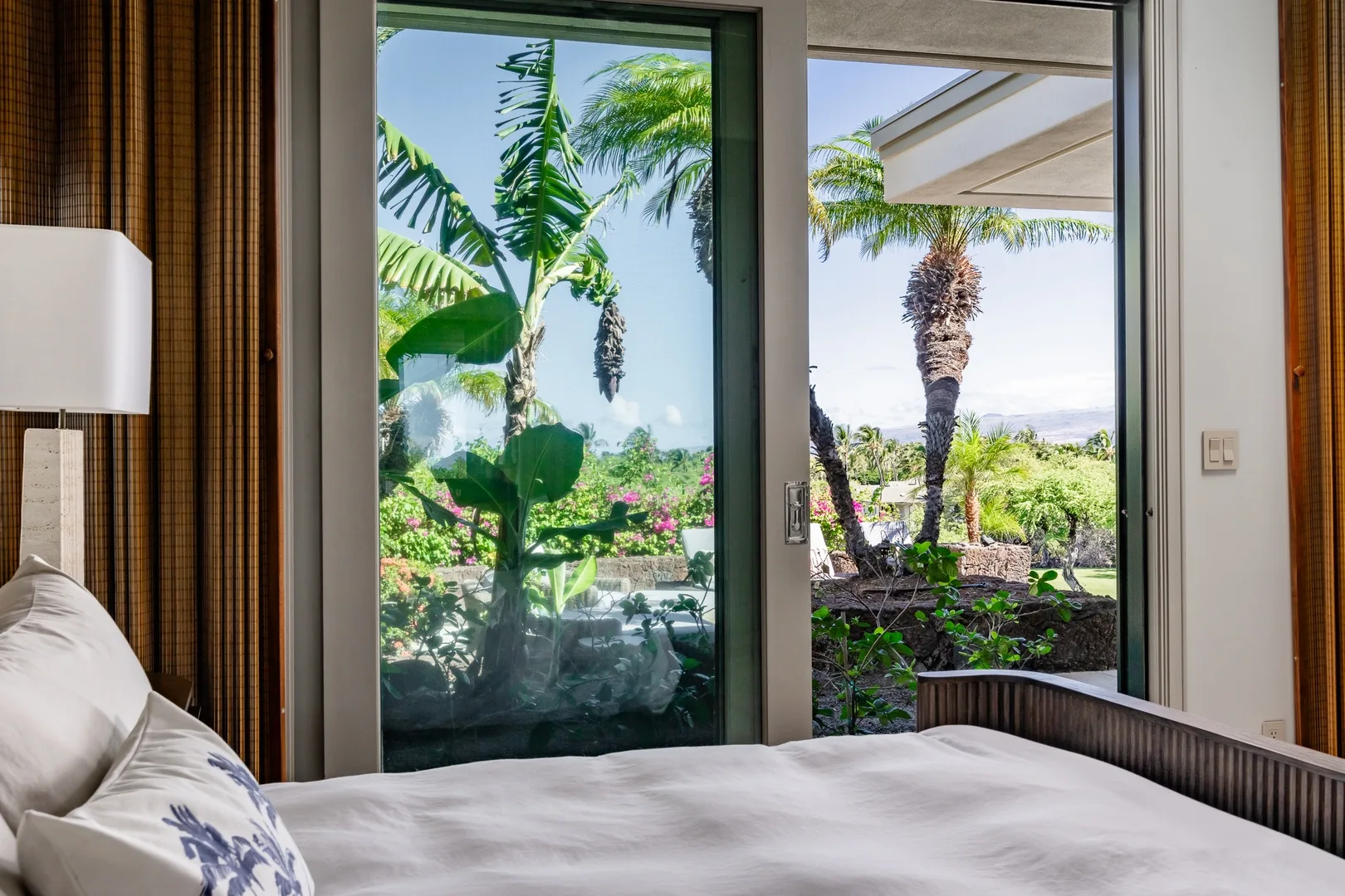 Un dormitorio con una cama junto a puertas de vidrio que dan a palmeras y vegetación.