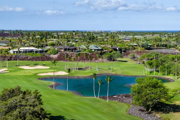 Un campo de golf ajardinado con un estanque, bunkers de arena y casas a lo lejos.
