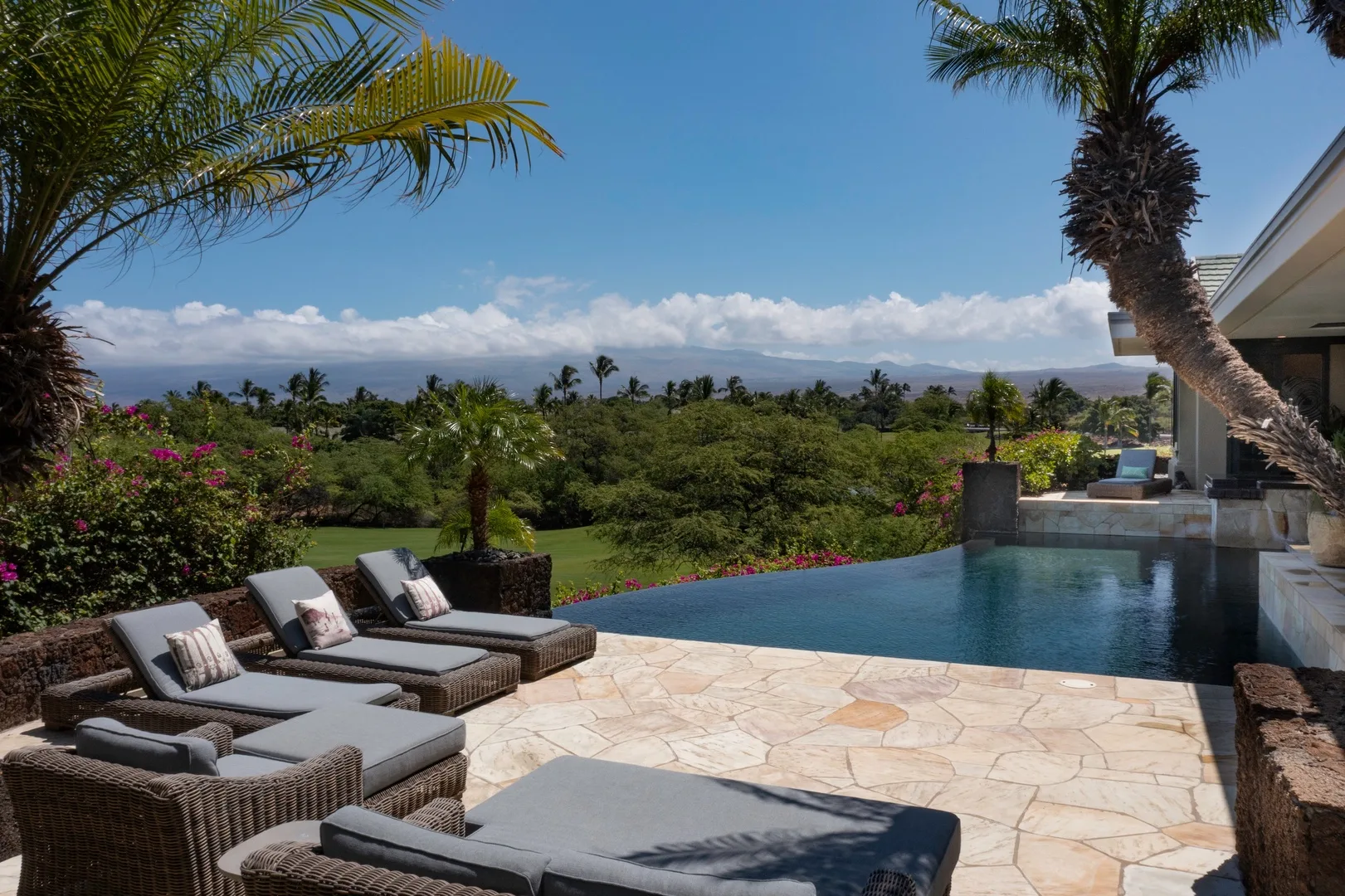 Un patio junto a una piscina exterior da a un jardín paisajístico, con asientos acolchados junto al agua.