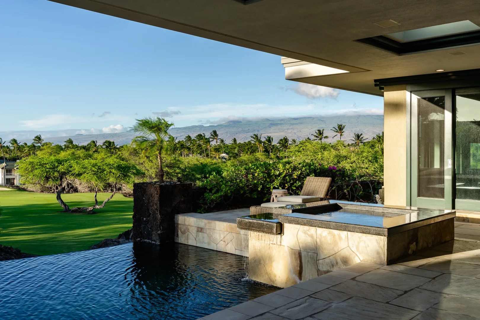 Una piscina exterior junto a un jacuzzi elevado, con césped y árboles al fondo.
