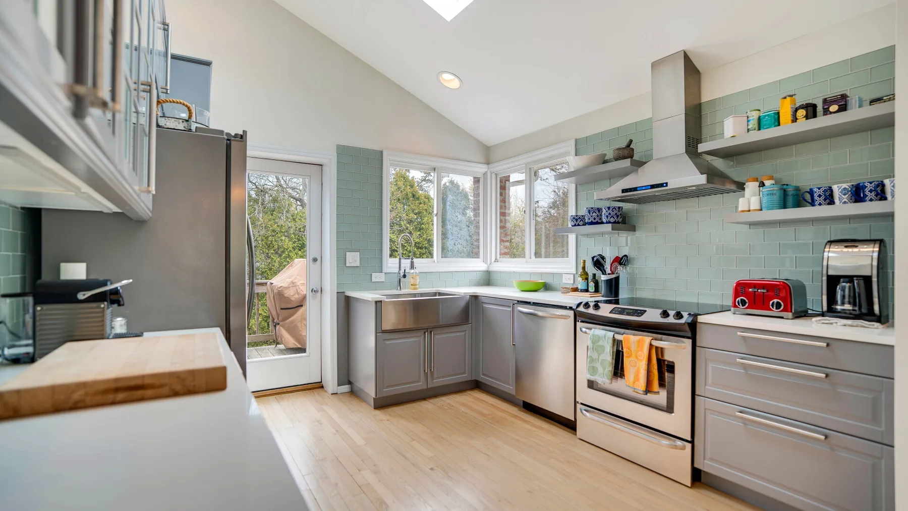 Una cocina luminosa con gabinetes grises, paredes de azulejos y ventanas grandes.