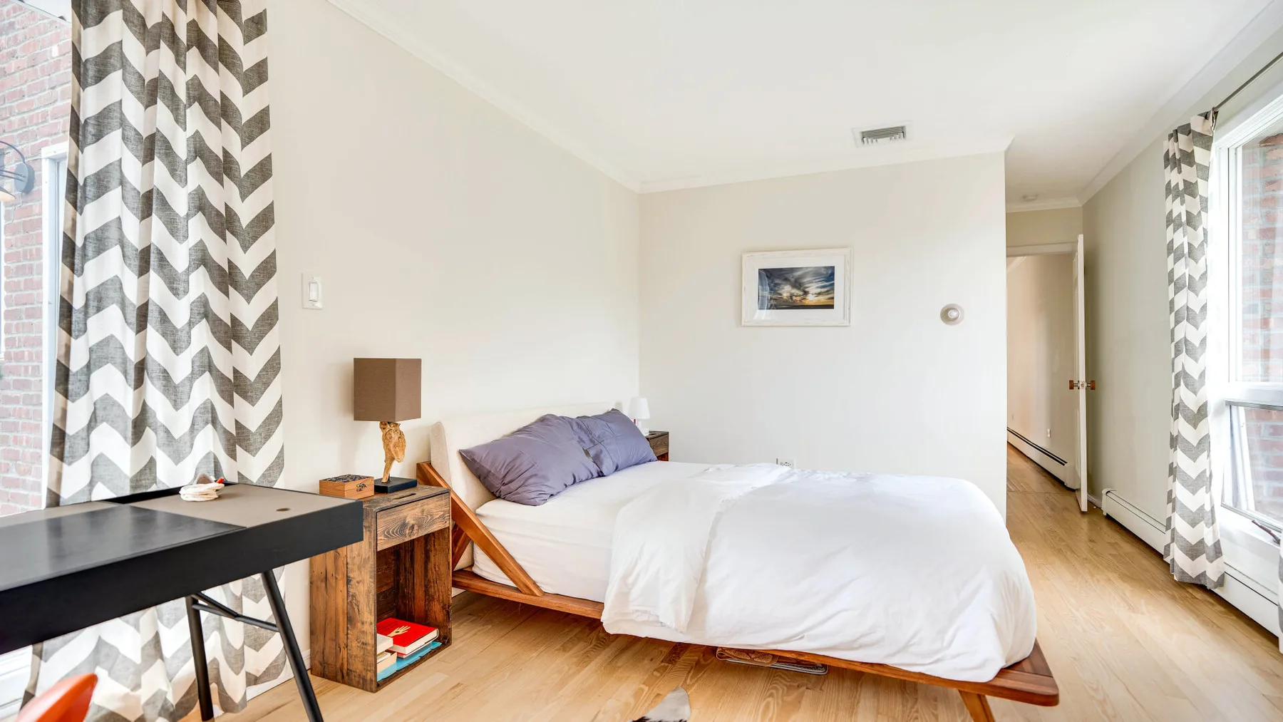 Un dormitorio luminoso con una cama, suelo de madera y ventanas grandes.
