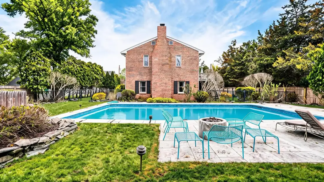 A backyard swimming pool sits beside a brick house, lawn, and outdoor seating.