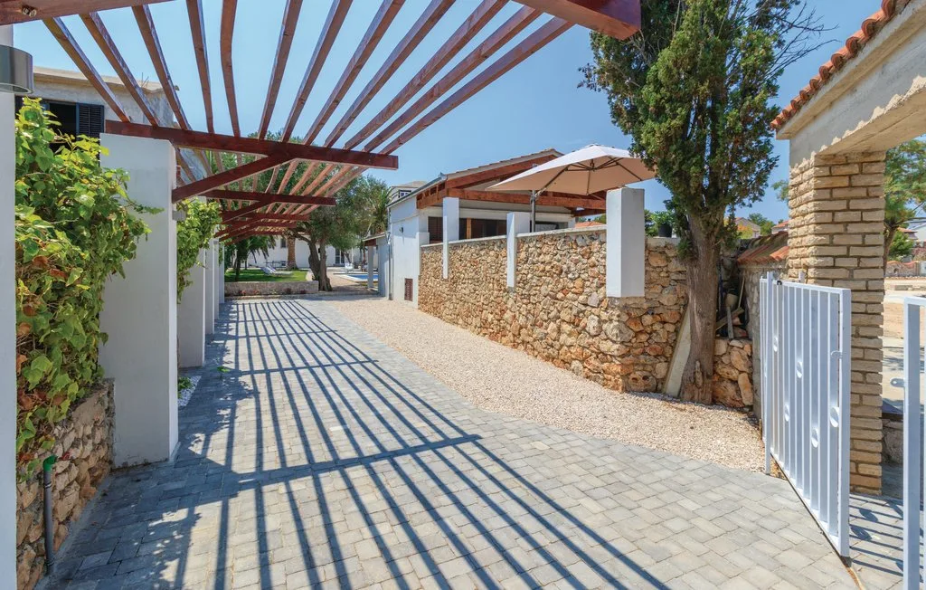 A paved outdoor walkway runs beneath a wooden pergola beside stone walls and greenery.