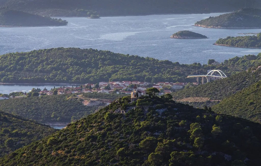A coastal landscape overlooks a town, wooded hills, and islands across the water.