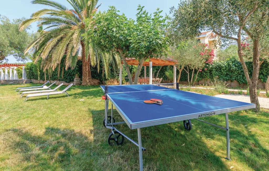 A ping-pong table sits in a grassy garden surrounded by trees and lounge chairs.