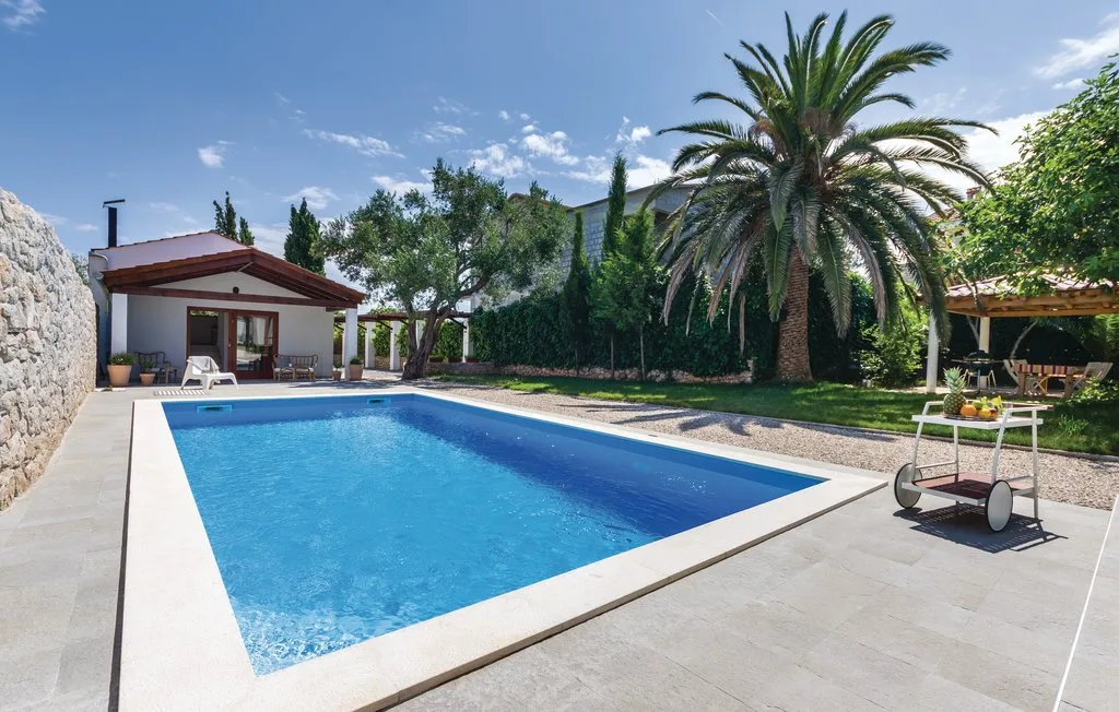 An outdoor swimming pool sits beside a patio with palm trees and greenery.
