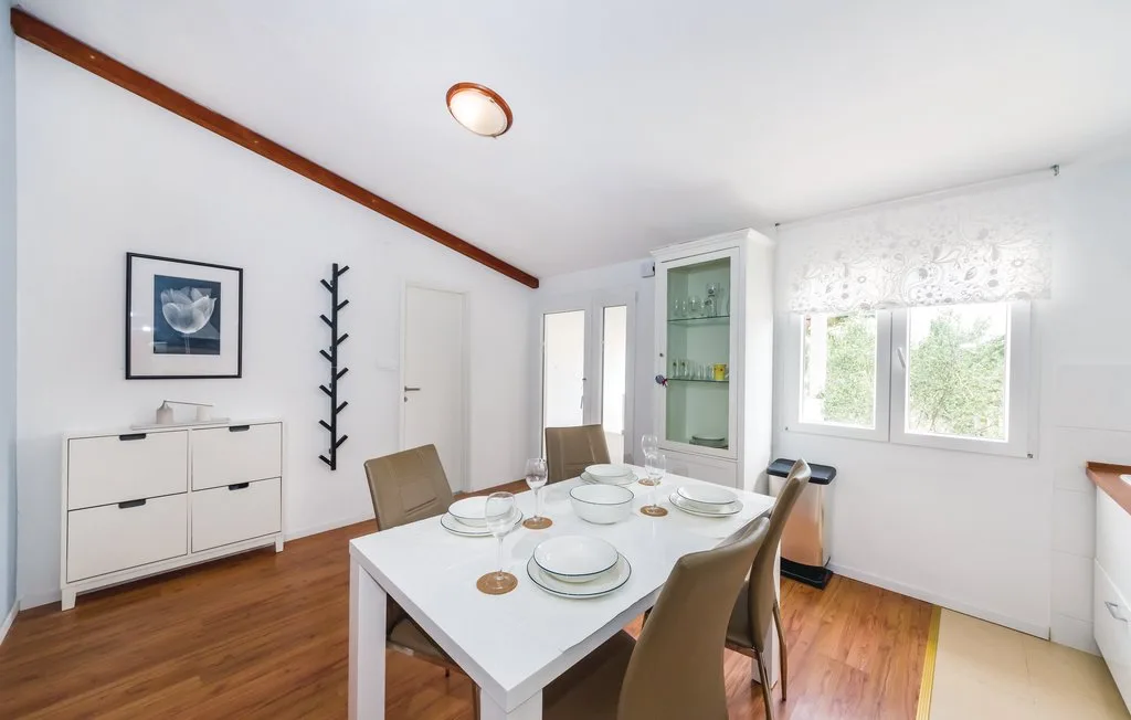 A bright dining area has a white table set beneath windows beside a glass-front cabinet.