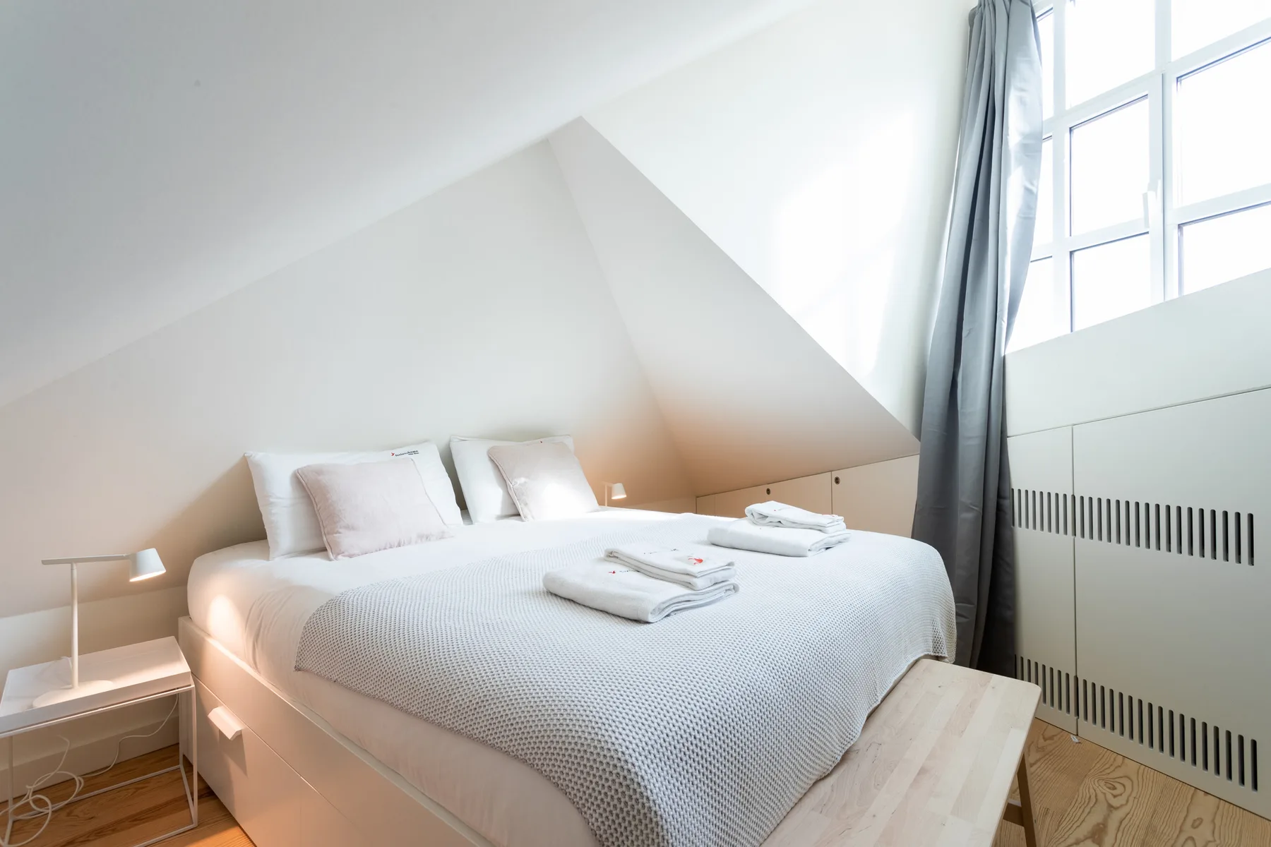 A bright bedroom with a neatly made bed beneath a sloped ceiling.
