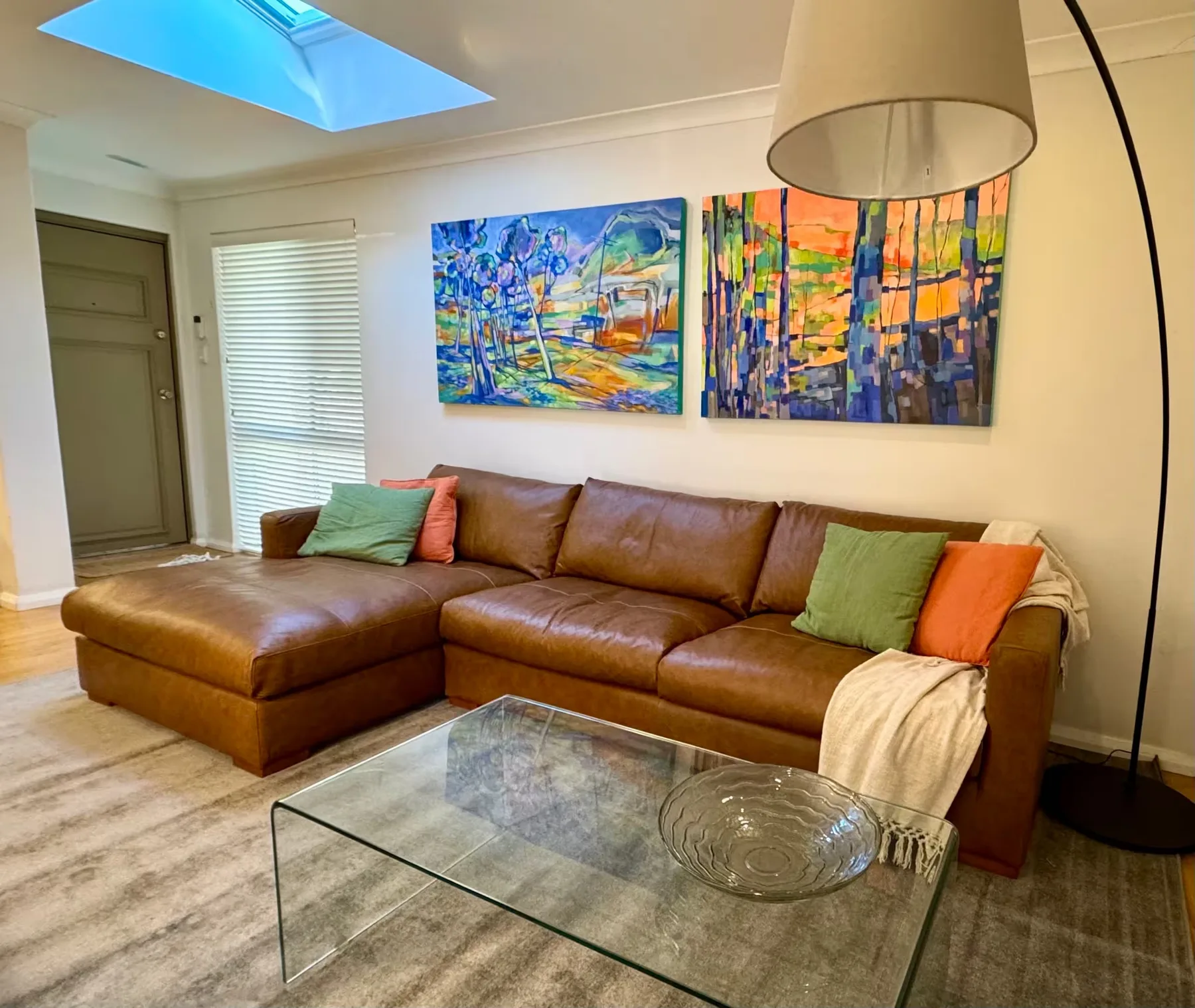 A living room with brown seating, colorful paintings, and a glass coffee table.
