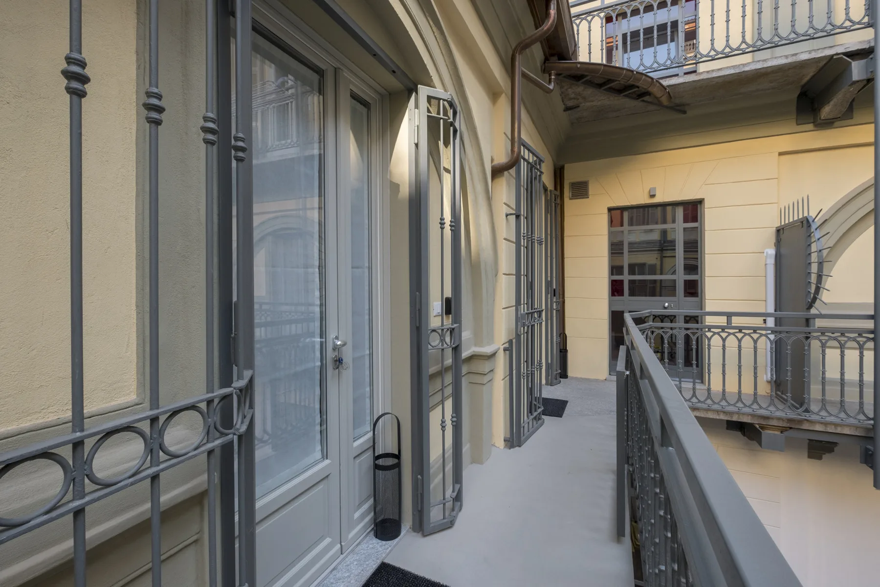 Un pasillo exterior de edificio con paredes claras y barandillas metálicas.
