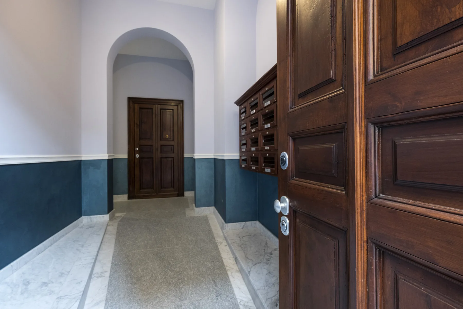 Un pasillo interior con puertas de madera oscura, buzones y paredes azules en la parte inferior.